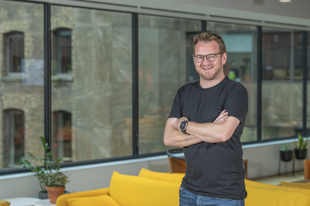 Dave Robbins, CEO and Co-Founder of Azarc stands in front of a large glass window, in Azarc's London office at Devonshire Square, in a room with a yellow sofa.